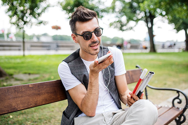 Giovane cieco con smartphone seduto su una panchina nel parco in città, chiamando.